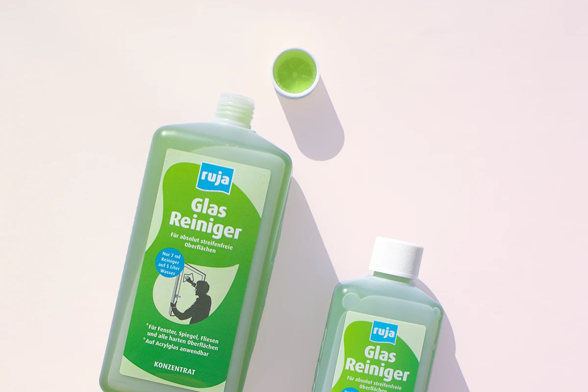 Zwei Flaschen ruja Glasreiniger mit gefülltem Deckel vor einem pastellfarbenen Hintergrund